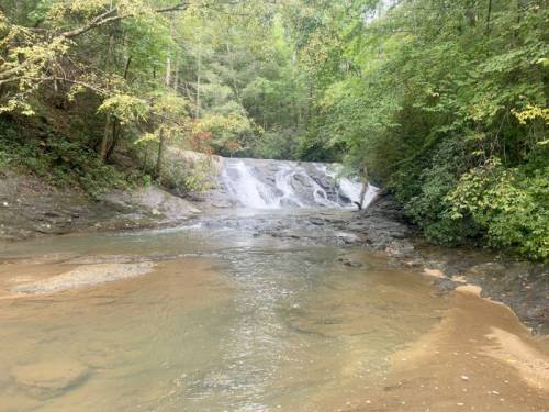 A serene waterfall cascading into a calm river, surrounded by lush green trees and rocky terrain.