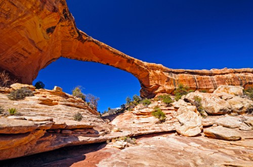 Natural rock arch against a clear blue sky, surrounded by rugged terrain and desert vegetation.
