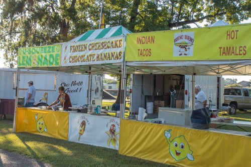 A colorful food stall at an outdoor event, offering lemonade, frozen cheesecake, chocolate bananas, and various snacks.