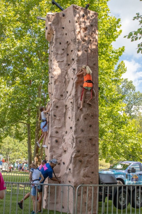 Children climbing a rock wall at an outdoor event, surrounded by trees and spectators.