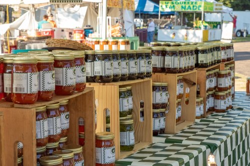 Jars of colorful preserves displayed on wooden stands at a market, with a checkered tablecloth in the foreground.