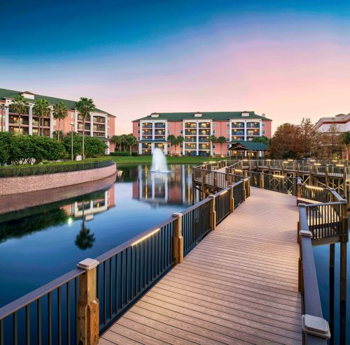 A wooden walkway leads to a serene lake with a fountain, surrounded by lush greenery and colorful buildings at sunset.