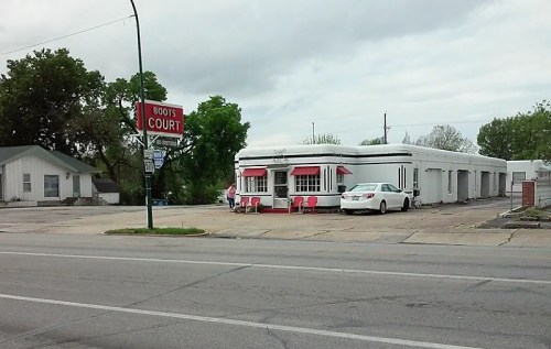 A vintage diner with red accents, surrounded by trees, and a white car parked outside on a cloudy day.