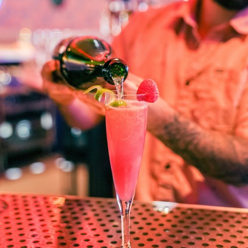 A bartender pours a drink into a glass garnished with a raspberry and lime, set against a vibrant bar background.