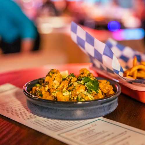 A bowl of orange cauliflower dish garnished with herbs, placed on a wooden table with a menu and fries in the background.