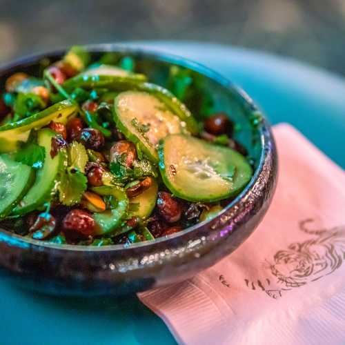 A vibrant salad with sliced cucumbers, herbs, and black beans in a dark bowl, resting on a pink napkin.