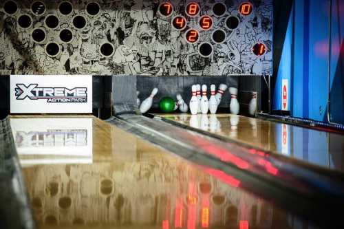 A bowling lane with pins set up, a green ball nearby, and a scoreboard displaying scores in the background.