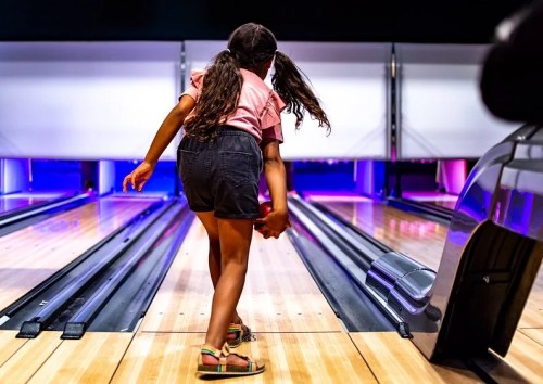 A young girl prepares to bowl, facing the lanes with a focused expression in a brightly lit bowling alley.