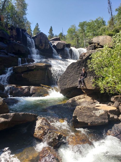 A person sits on a rock by a waterfall, surrounded by lush greenery and flowing water under a clear blue sky.