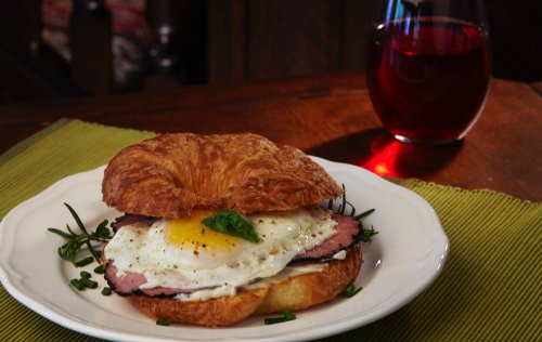A croissant sandwich with ham, a fried egg, and herbs, served on a plate with a glass of red drink in the background.