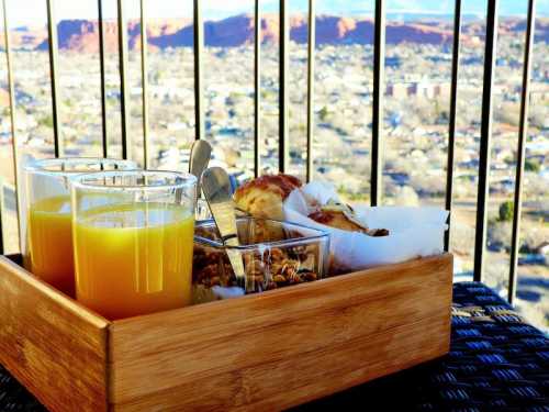 A wooden tray with two glasses of orange juice, granola, and pastries, set against a scenic view of a landscape.