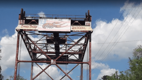 A rusted bridge structure with a large banner reading "Breaux Bridge, Louisiana" against a blue sky with clouds.