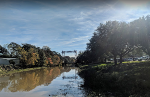 A calm river reflects trees and a bridge under a clear sky, with sunlight filtering through the branches.