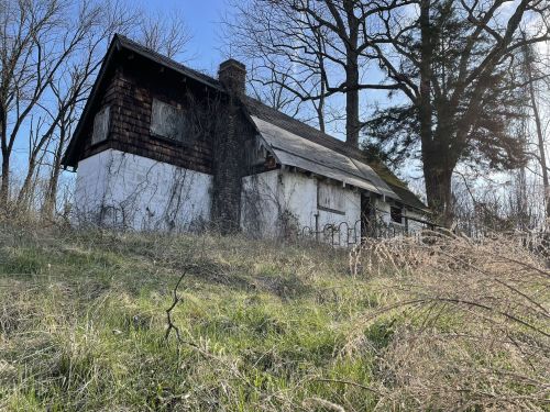 An abandoned house with a weathered exterior, surrounded by tall grass and trees under a clear blue sky.