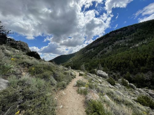 A winding dirt path through a mountainous landscape, surrounded by greenery and under a partly cloudy sky.