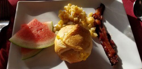 A plate with watermelon slices, diced potatoes, a muffin, and crispy bacon.