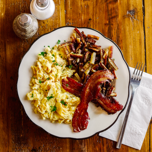 A plate of scrambled eggs, crispy bacon, and golden-brown hash browns on a wooden table. Salt and pepper shakers nearby.