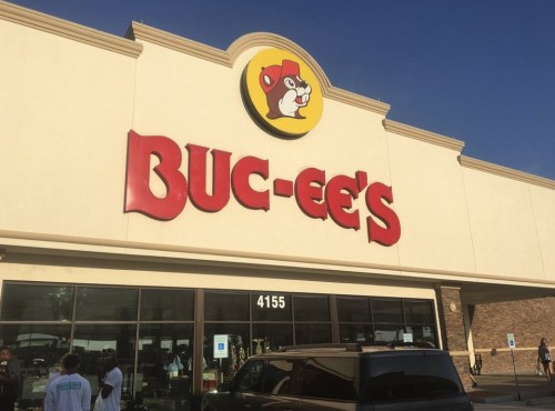 Exterior of a Buc-ee's convenience store with a large logo and bright blue sky. People are gathered outside.