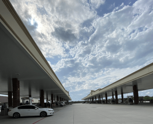 A wide view of a gas station canopy with cars parked underneath, set against a cloudy sky.