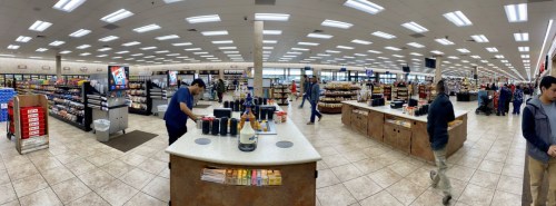 A spacious convenience store with shelves of snacks, drinks, and a busy checkout area. Customers browse and shop.