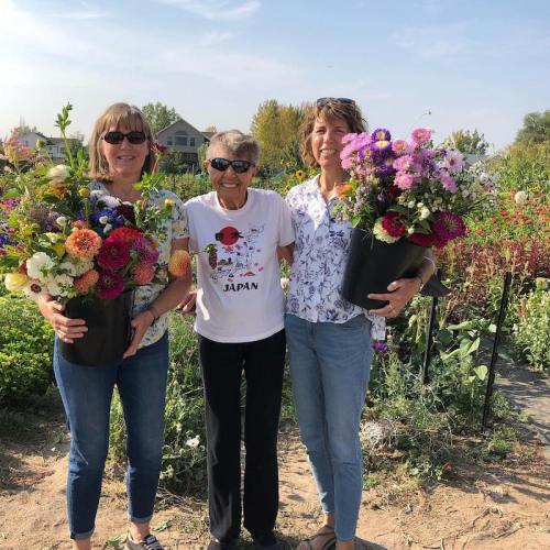 Three women stand together in a garden, each holding a pot of colorful flowers, smiling at the camera.
