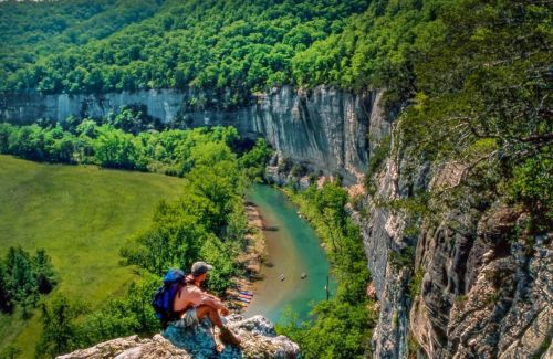 A hiker sits on a rocky ledge overlooking a lush green valley and winding river below.