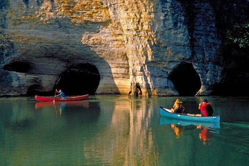Two kayakers paddle on a calm river near rocky cliffs, with one person taking a photo of the scenery.