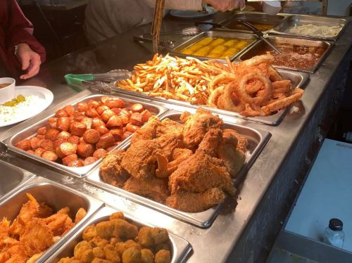 A buffet spread featuring fried chicken, sausages, fries, onion rings, and various side dishes in metal trays.