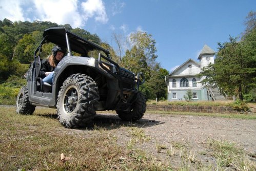 A person drives an ATV near an old, abandoned building surrounded by trees and blue sky.