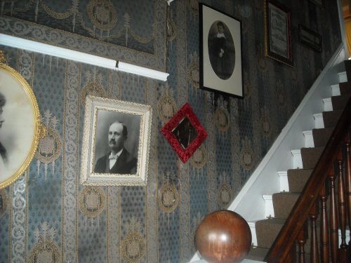 A staircase with patterned wallpaper, featuring various framed portraits and a decorative wooden sphere.