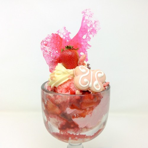 A colorful dessert in a glass, featuring strawberry ice cream, whipped cream, a strawberry, and decorative pink elements.