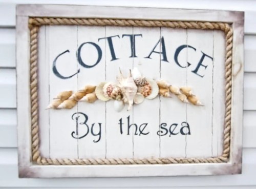 A decorative sign reading "Cottage By the Sea," adorned with seashells and rope accents.