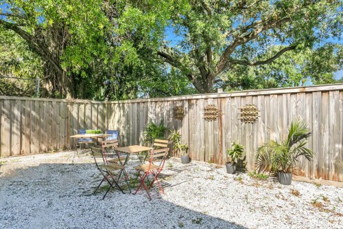 A cozy outdoor space with a gravel floor, wooden fence, and potted plants, featuring a small table and chairs.