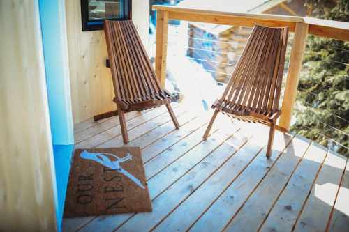 Two wooden chairs on a balcony with a welcome mat that says "OUR NEST," surrounded by a serene outdoor setting.