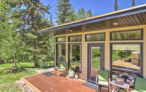 A modern cabin exterior with large windows, a wooden deck, and green chairs, surrounded by trees and grass.