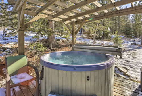 A wooden hot tub under a pergola, surrounded by snow-covered trees and a wooden deck.