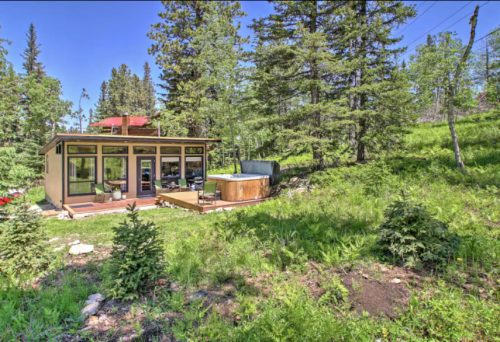 A modern cabin surrounded by trees, featuring a deck and hot tub in a lush green landscape.