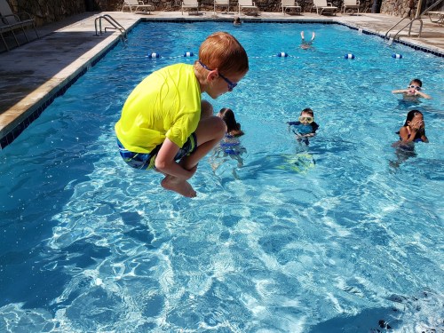 A child in a bright yellow shirt jumps into a pool, while other kids swim nearby.