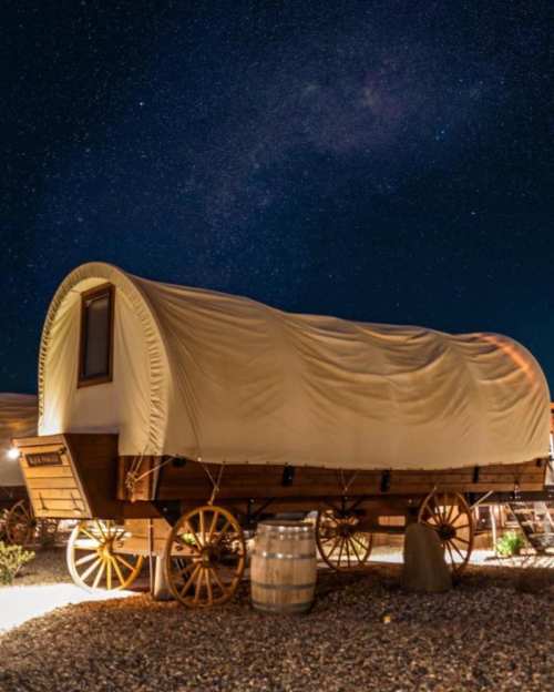 A covered wagon under a starry night sky, surrounded by barrels and gravel.