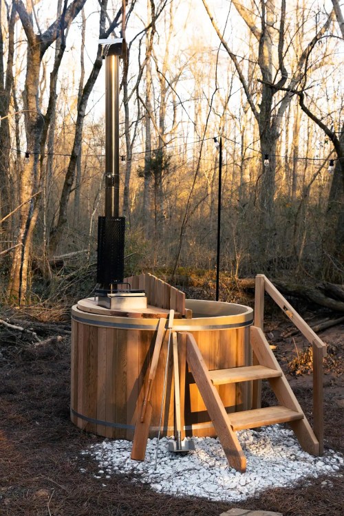 A wooden hot tub with a chimney, surrounded by trees and gravel, in a serene outdoor setting.