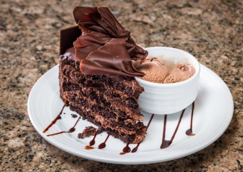 A slice of chocolate cake topped with chocolate shavings, served with a scoop of chocolate ice cream on a white plate.