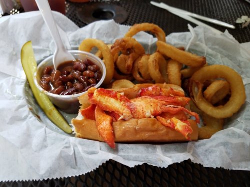 A lobster roll topped with lobster meat, served with onion rings, baked beans, and a pickle on a paper-lined plate.