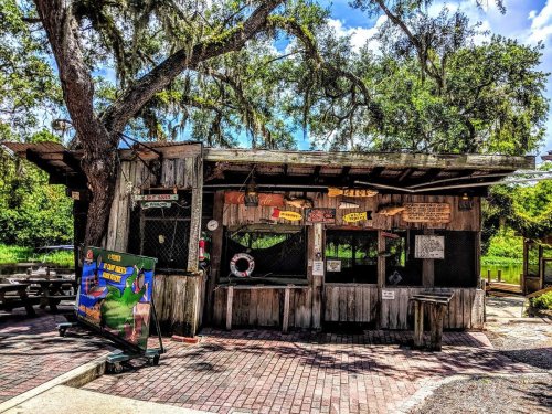 A rustic wooden shack with a sign and picnic tables, surrounded by trees and greenery on a sunny day.