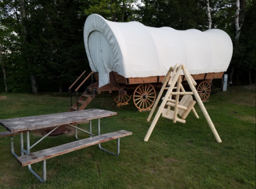 A covered wagon on wheels beside a wooden picnic table and swing set in a grassy area surrounded by trees.