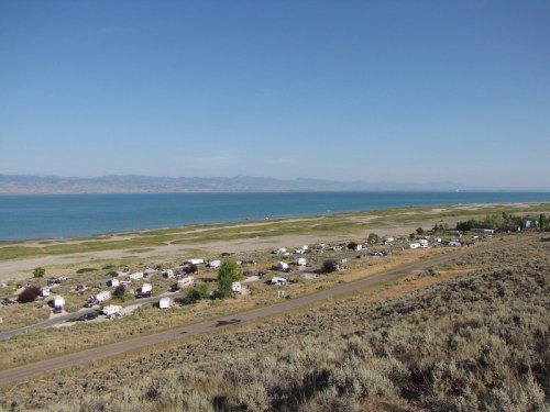 A scenic view of a lake with a shoreline, surrounded by grassy areas and a cluster of RVs in the foreground.