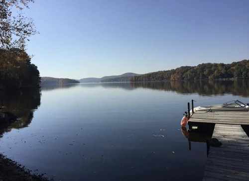A calm lake reflects the clear sky, with a wooden dock on the shore and autumn foliage in the background.