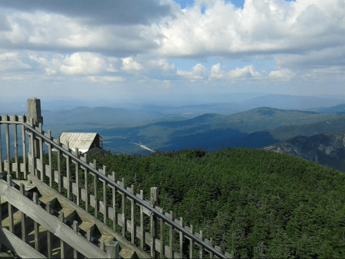 A scenic view from a mountain top, featuring lush green trees, a wooden railing, and distant blue hills under a cloudy sky.
