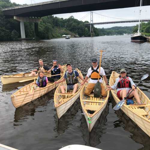 A group of six people in kayaks on a river, with a bridge and trees in the background.