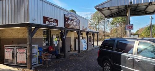 A small roadside cafe with a sign reading "Canyon Cafe," featuring outdoor seating and a nearby gas station.