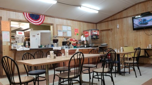 A cozy diner interior with wooden walls, tables, chairs, a soda machine, and a TV displaying a blue airplane.